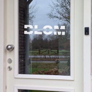 Een voordeur met op het glas een opdruk van de achternaam onderbroken door een horizontale lijn met daarop de voornamen in sierlijk lettertype