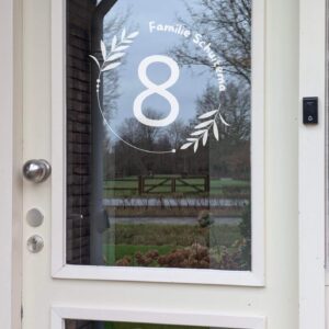 Een voordeur met op het glas een opdruk van een cirkel met blaadjes onderbroken door de naam en in het midden het huisnummer