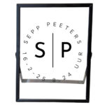 Een zwarte fotolijst met een cirkel gevormd door de geboortegegevens; naam, datum en tijd. In het midden van de cirkel staan de initialen.