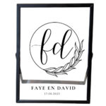 Een zwarte fotolijst met daarin een sierlijke cirkel met blaadjes. In de cirkel staan 2 initialen en onder de cirkel staan 2 namen met de datum.