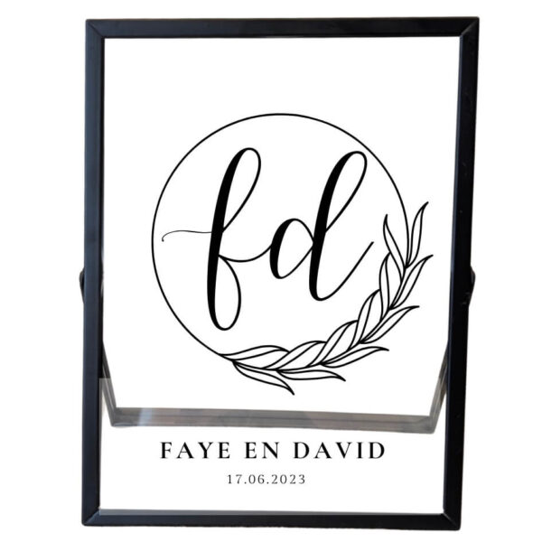 Een zwarte fotolijst met daarin een sierlijke cirkel met blaadjes. In de cirkel staan 2 initialen en onder de cirkel staan 2 namen met de datum.