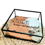 Vierkant glazen kistje met een zwart frame en bedrukking van een omlijnde naam met daaronder in sierlijke letters: haar eerste pakje.