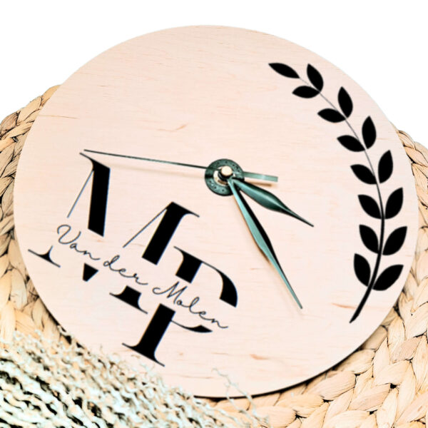 Een blanco houten ronde klok met zwarte wijzers en een opdruk van een sierlijke tak met links onder de initialen met daarin de achternaam.