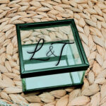 Vierkant glazen kistje met zwart frame en een opdruk van de twee initialen met een en teken er tussen in sierlijk lettertype geschreven