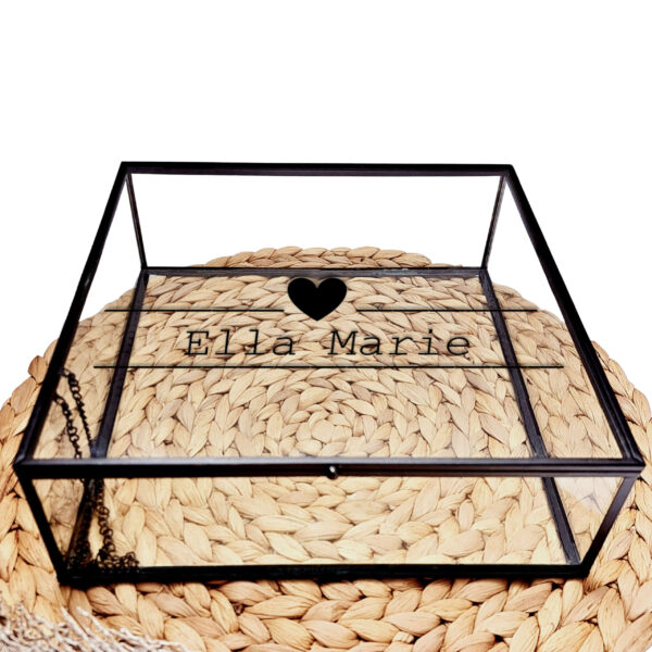 Glazen kistje met zwart frame en een zwarte opdruk van de naam tussen twee lijnen waarvan de bovenste is onderbroken door een hartje