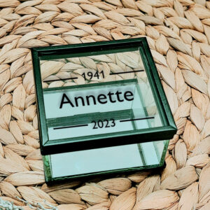 Vierkant glazen kistje met zwart frame en een opdruk van een naam met daarboven en onder een lijn welke onderbroken is door een jaartal