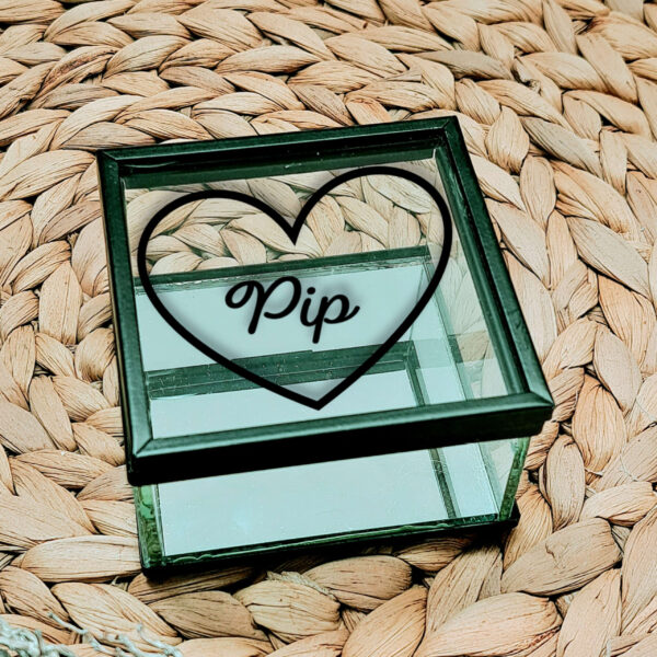 Glazen kistje met zwart frame en een zwarte opdruk van hartje met daarin een naam in sierlijke letters geschreven.