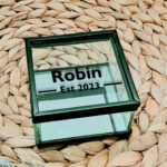 Glazen kistje met zwart frame en een zwarte opdruk van de naam met daaronder een lijn welke onderbroken wordt door de tekst: Est plus jaartal.