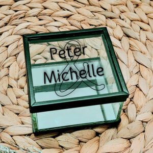 klein vierkant glazen kistje met zwart frame en een opdruk van twee namen onder elkaar met daarachter de omlijning van een en teken.