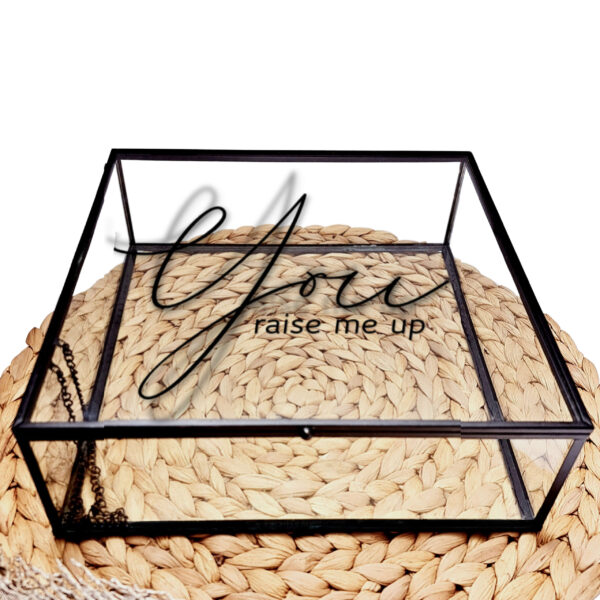 Glazen kistje met zwart frame en een zwarte opdruk van de tekst: you raise me up, waarvan you in een sierlijk lettertype staat.