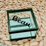 Glazen kistje met zwart frame en een zwarte opdruk van een naam in sierlijke letters geschreven.