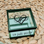 Vierkant glazen kistje met zwart frame en een opdruk van een sierlijk hartje, aan de voorkant van het kistje staat een datum gedrukt.