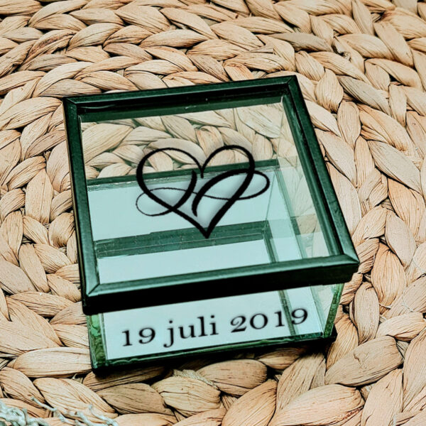 Vierkant glazen kistje met zwart frame en een opdruk van een sierlijk hartje, aan de voorkant van het kistje staat een datum gedrukt.