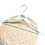 Een wit houten kleerhanger met daarop de bedrukking van de naam in sierlijke letters, links de rol en rechts de datum.