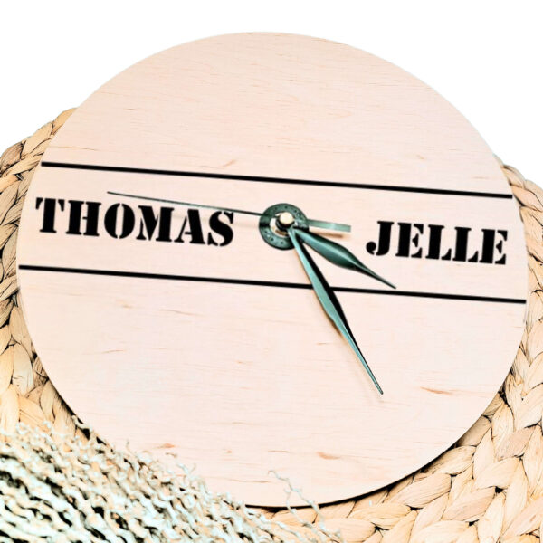 Een blanco houten ronde klok met zwarte wijzers en een opdruk van twee namen in stoere letters met daar boven en onder een lijn.