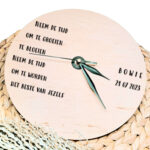 Een blanco houten ronde klok met zwarte wijzers en een opdruk van een gedicht aan de linkerkant en de naam met datum aan de rechterkant.