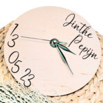 Een blanco houten ronde klok met zwarte wijzers en een opdruk van de namen rechts bovenin en de datum links onderin.