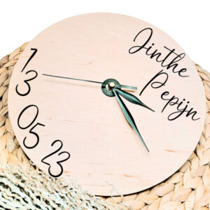 Een blanco houten ronde klok met zwarte wijzers en een opdruk van de namen rechts bovenin en de datum links onderin.