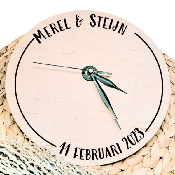 Een blanco houten ronde klok met zwarte wijzers en een opdruk van een cirkel welke links onderbroken is door de namen en rechts door de datum.