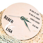 Een blanco houten ronde klok met zwarte wijzers en een opdruk van een gedicht rechts boven en de namen links onder met een en teken erachter