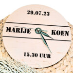 Een blanco houten ronde klok met zwarte wijzers en een opdruk van twee namen tussen 2 lijnen met daarboven de datum en daaronder de tijd.