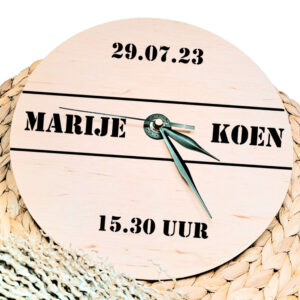 Een blanco houten ronde klok met zwarte wijzers en een opdruk van twee namen tussen 2 lijnen met daarboven de datum en daaronder de tijd.