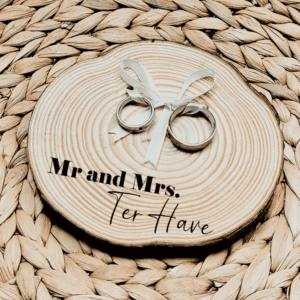 Rond ringplankje gemaakt van een boomstamschijf met daarop 2 ringen aan satijnen lint en een zwarte tekst: mr and mrs met sierlijke achternaam