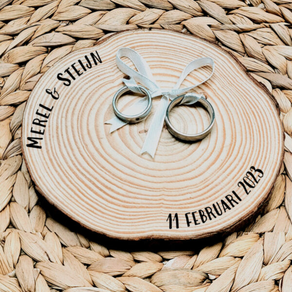 Rond ringplankje gemaakt van een boomstamschijf met daarop 2 ringen aan een satijnen lint en een zwarte opdruk van de namen en datum.