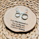 Rond ringplankje gemaakt van een boomstamschijf met daarop ringen aan satijnen lint en een zwarte tekst uit het lied: from this moment on.