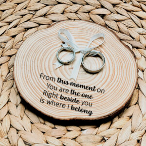 Rond ringplankje gemaakt van een boomstamschijf met daarop ringen aan satijnen lint en een zwarte tekst uit het lied: from this moment on.