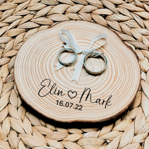 Rond ringplankje gemaakt van een boomstamschijf met daarop ringen aan satijnen lint en een zwarte opdruk van namen met een hartje en de datum