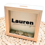 Een blanco houten vierkante spaarpot waarvan het glas bedrukt is met de naam en daaronder in sierlijke dunne letters; Zijn/Haar eerste miljoen