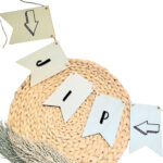 Blanco houten vlaggetjes aan een jute touw met op elke vlag een zwarte letter die samen een naam vormen en op de buitenste vlaggen een pijl.