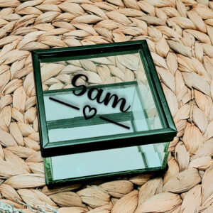 Glazen kistje met zwart frame en een diagonale opdruk van de naam met daaronder een lijn welke onderbroken wordt door een hartje.