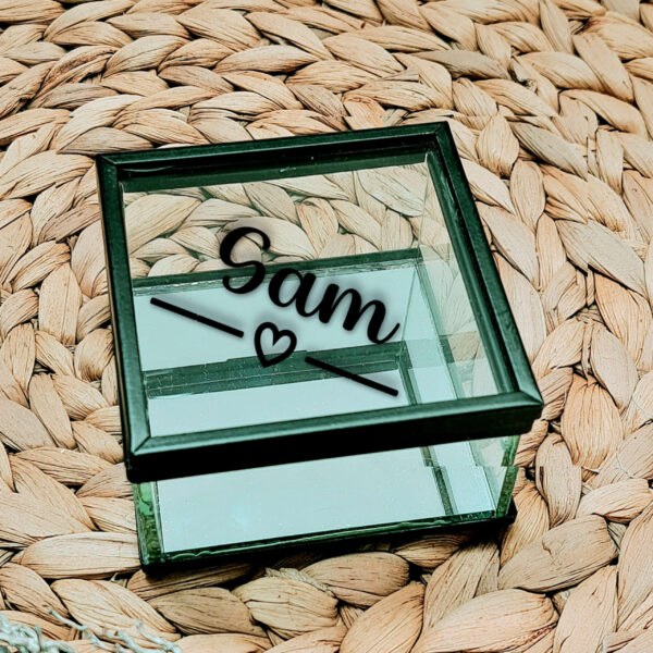 Glazen kistje met zwart frame en een diagonale opdruk van de naam met daaronder een lijn welke onderbroken wordt door een hartje.